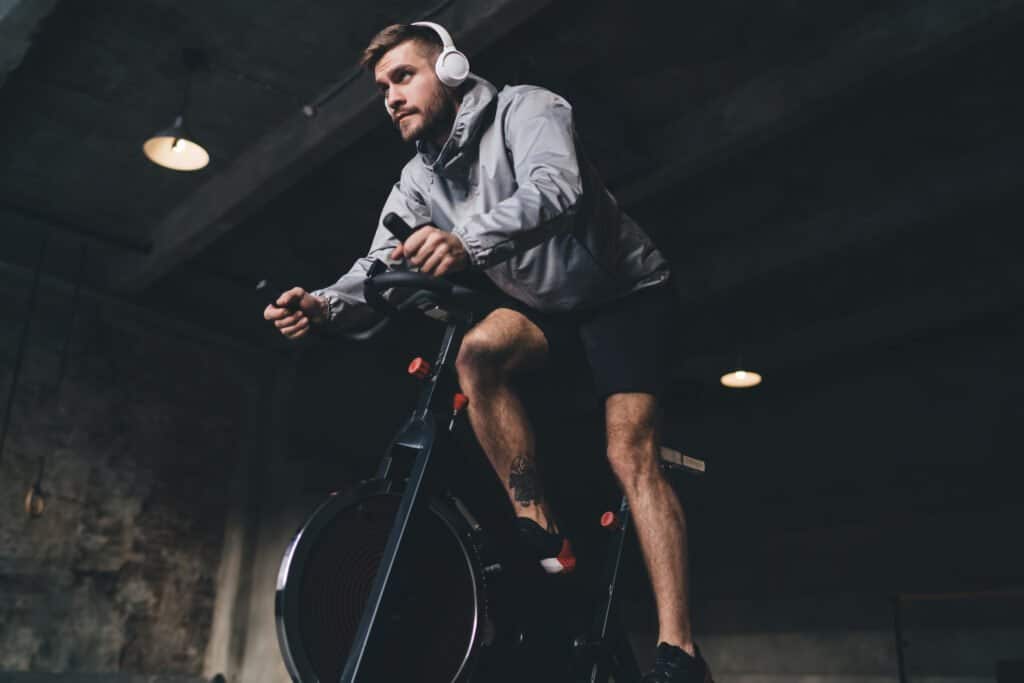 Focused young man training on a stationary bike indoors, wearing headphones, sports attire, and a heart rate monitor.