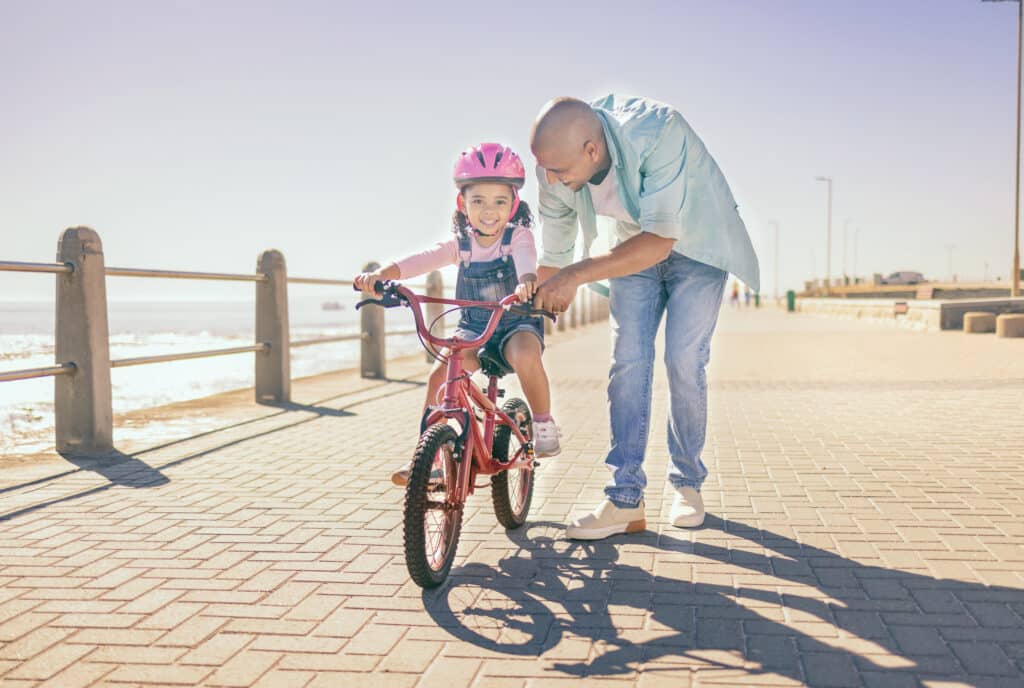 Father helping his daughter ride a bicycle on a sunny day, with the ocean visible in the background.