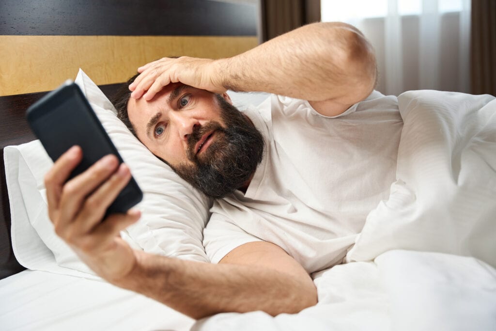 Man up late and cannot fall asleep. Looking at his phone thinking of restful sleep.