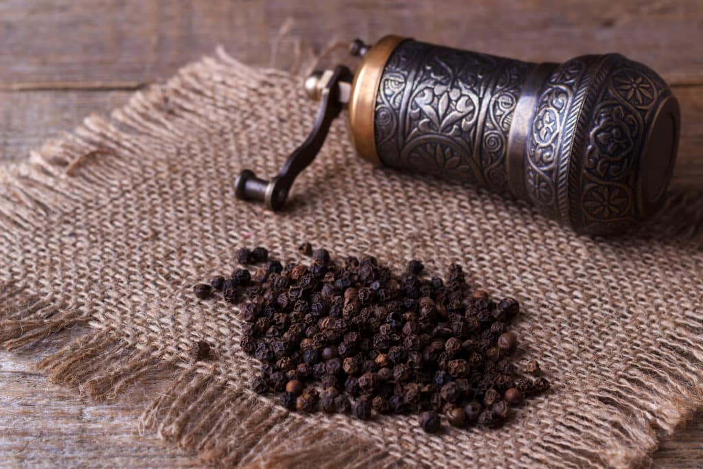 Black peppercorns on a woven jute surface with a traditional ornate metal pepper grinder beside them.