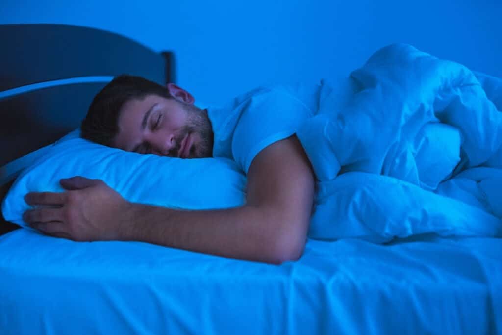 Man peacefully sleeping in bed under a blue light, wrapped in a blanket with a relaxed expression. - was melatonin his trick?