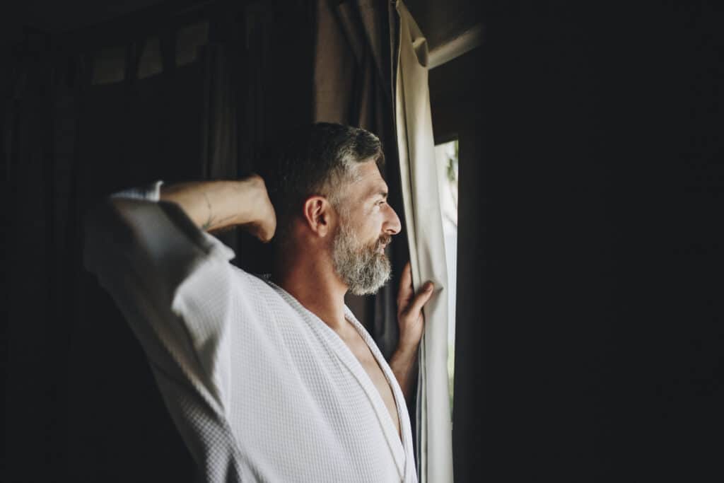 a man in a robe looking out a window thinking about melanin vs melatonin