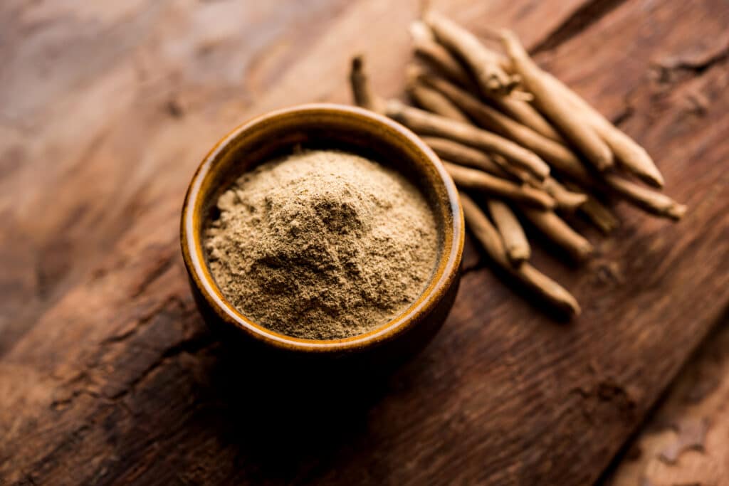 A wooden bowl with Ashwagandha powder next to Ashwagandha roots, all on an aged wooden table. Ashwagandha is known as one of the best supplements for sleep and anxiety reduction.