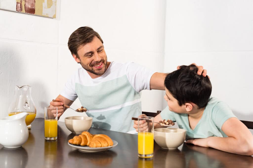 Smiling father with son at breakfast table enjoying orange juice and cereal. Dad is enjoying optimal health with Vitamin D3 and Zinc benefits.