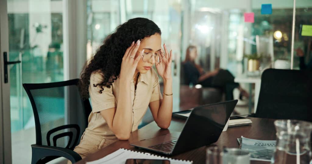 A woman at work with elbows on the desk and rubbing her temples and looking tired. What can help with stress reduction management.