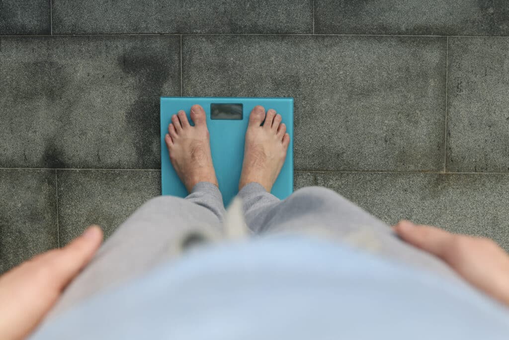 A heavier man standing on a scale with perception from the view of him looking down at increased weight, wondering How Hormones affect wright loss.