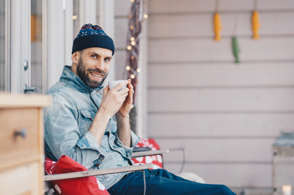 Positive glad attractive male model holds cup of tea, has good rest at balcony, breathes fresh air and enjoys sunny weather, has pleasant smile on face, feels delighted and pleased. Calm Your Nerves article for Men by Adaptafy.