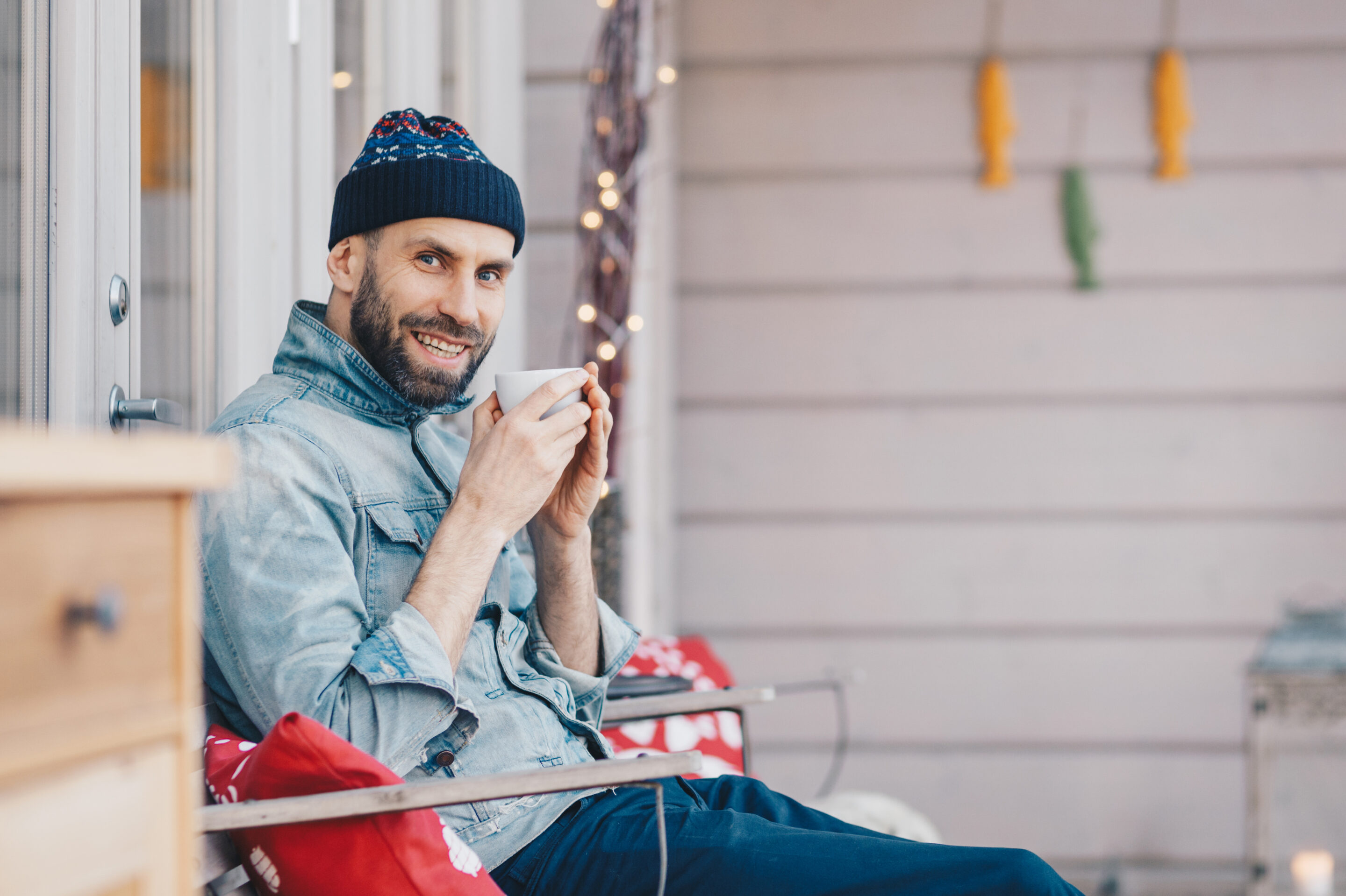 Positive glad attractive male model holds cup of tea, has good rest at balcony, breathes fresh air and enjoys sunny weather, has pleasant smile on face, feels delighted and pleased. Calm Your Nerves article for Men by Adaptafy.