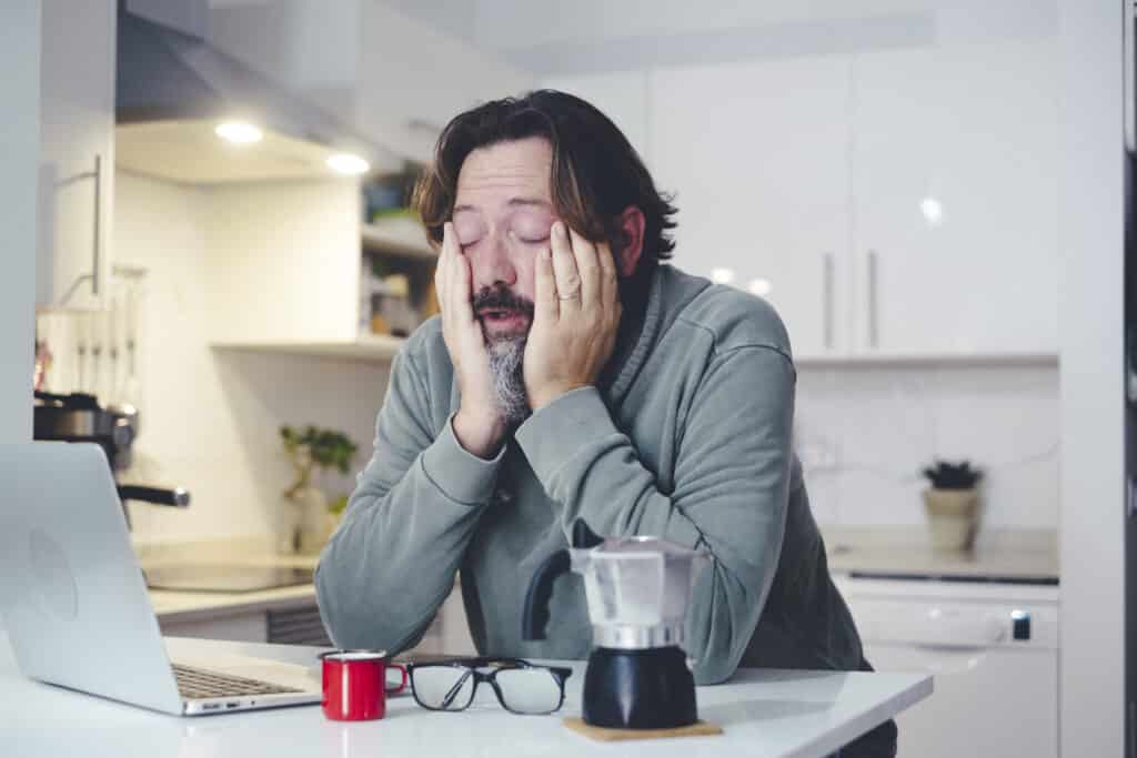 Tired man at home in morning wake up activity touching face and stretching after bad rest night. Coffee and computer online work concept. One person and insomnia health condition. Before job activity. Sleep Recovery Systems for Busy Fathers article by Adaptafy.