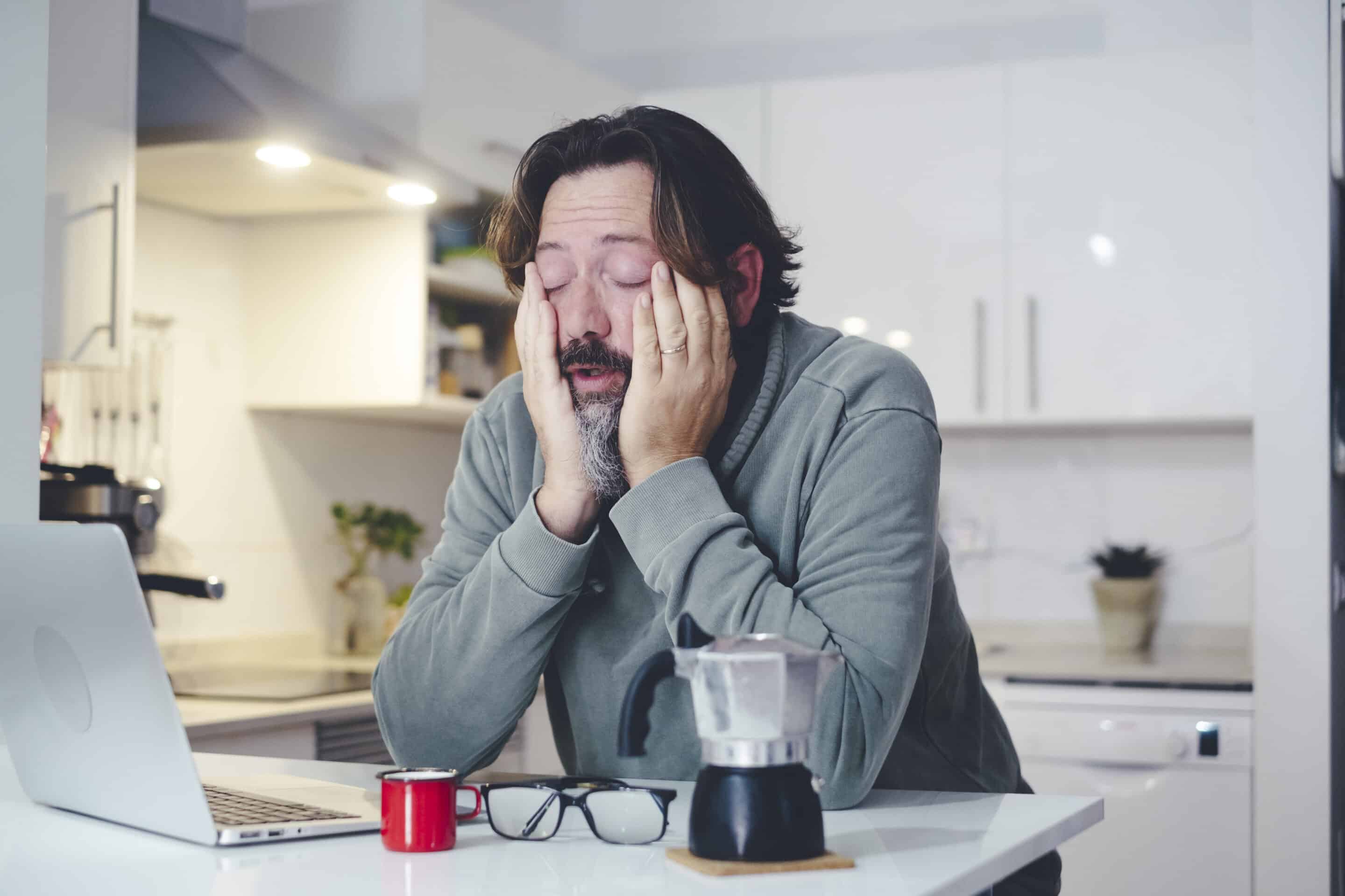 Tired man at home in morning wake up activity touching face and stretching after bad rest night. Coffee and computer online work concept. One person and insomnia health condition. Before job activity. Sleep Recovery Systems for Busy Fathers article by Adaptafy.